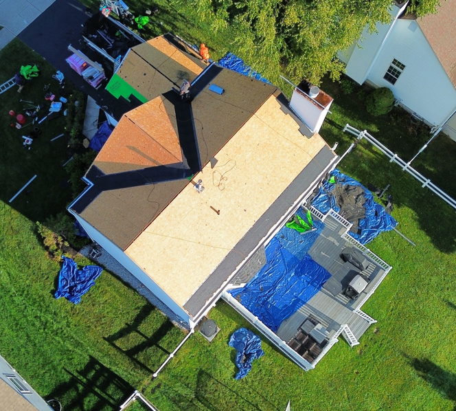 Aerial view of a house roof and backyard with blue tarps and scattered construction materials on grass.
