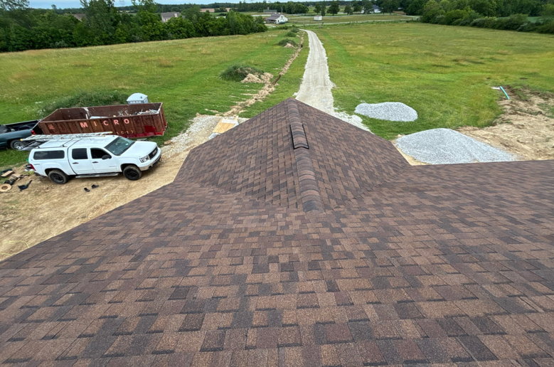 Aerial view of a brown shingle roof, driveway, white SUV, and open grassy lot with a gravel road.