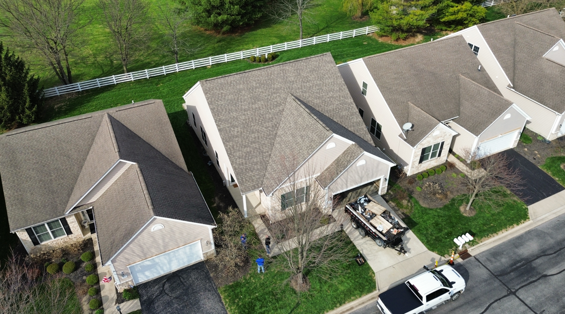 Aerial view of suburban homes with gray roofs, front yards, and a street with parked cars.