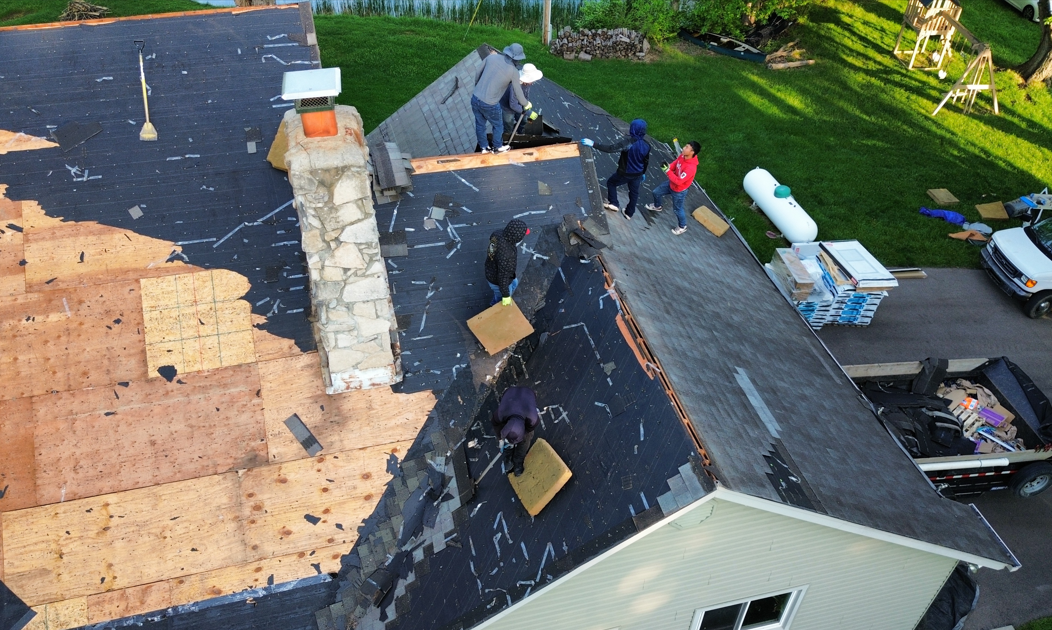 Roofers repairing a black shingle roof beside a brick chimney and exposed plywood deck