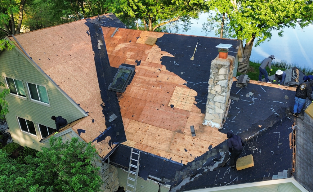 Aerial view of a house roof under repair, with exposed plywood, black underlayment, and a stone chimney.