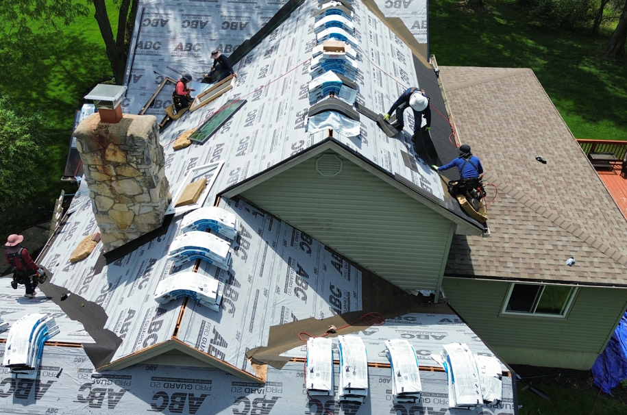 Roofing crew replacing shingles on a house roof with visible underlayment and ladders.