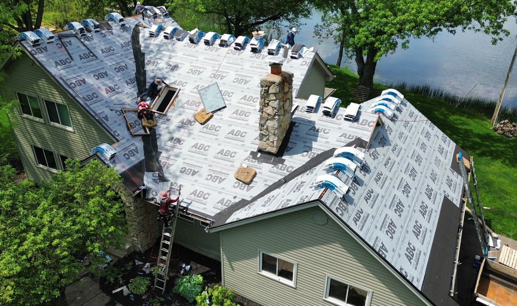 Aerial view of a house with a new gray roof, roofers working, and trees by a river.
