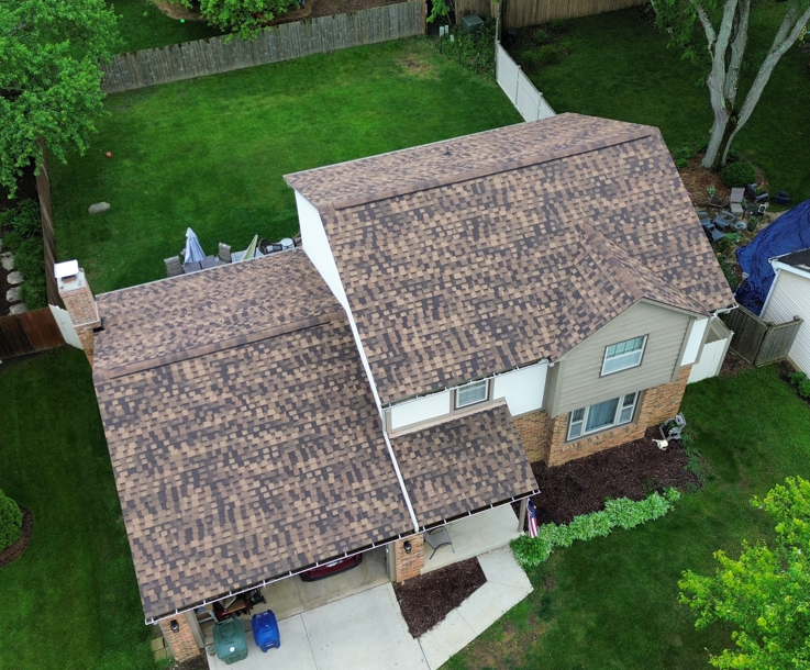 Aerial view of a tan house with brown shingle roof, attached garage, and green backyard
