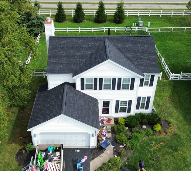 Aerial view of a white two-story house with a black roof, garage, and fenced green yard.