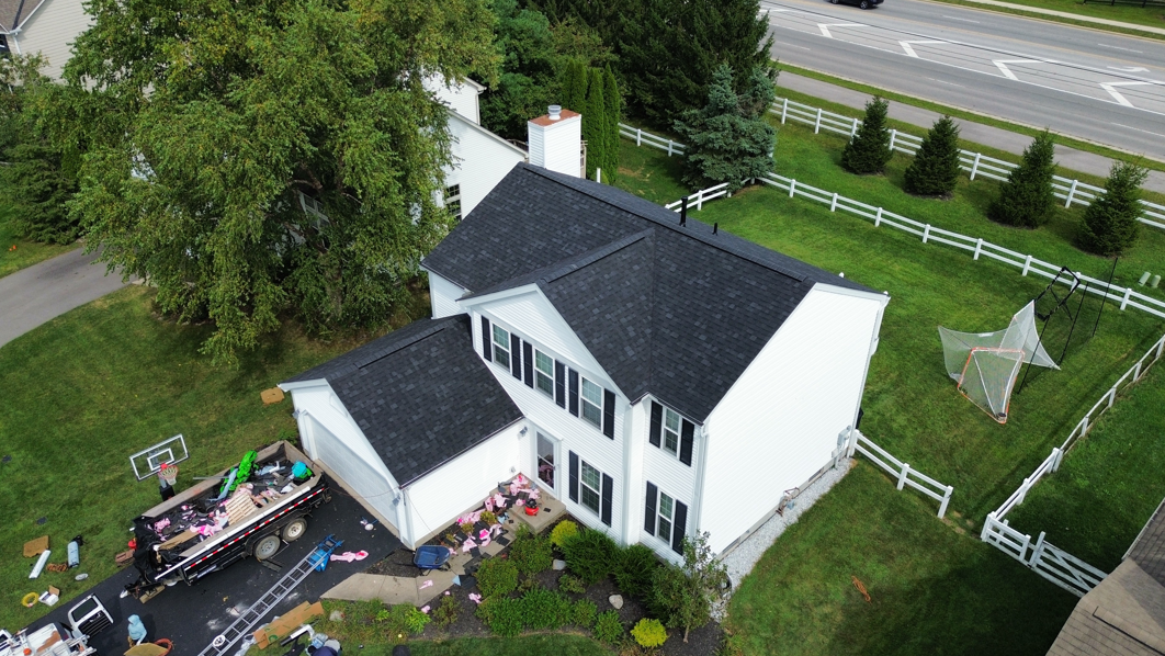Aerial view of a white house with a dark roof, yard, patio items, and a road behind it.