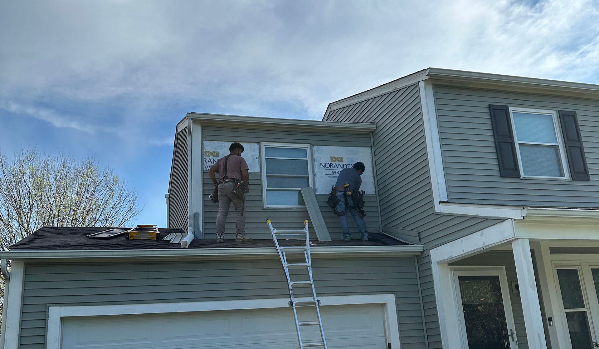 Workers on a rooftop using a ladder beside a two-story house with gray siding.