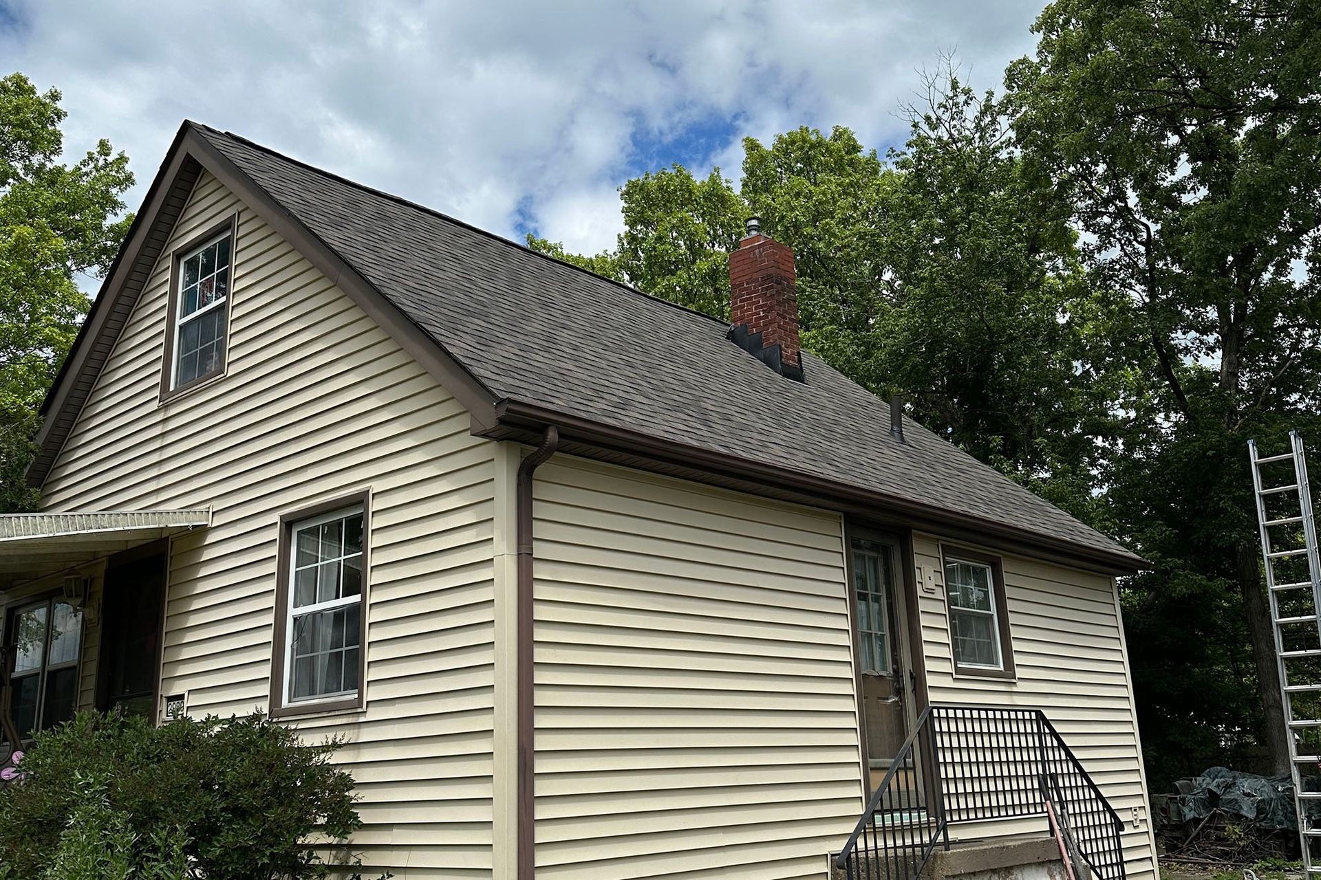 Small beige house with gray roof and white-trimmed windows, seen from the side with trees and a ladder nearby