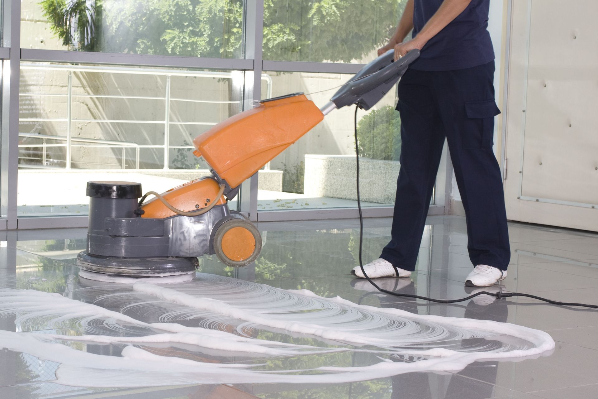 Person cleaning a floor with an orange floor scrubber in a bright indoor space
