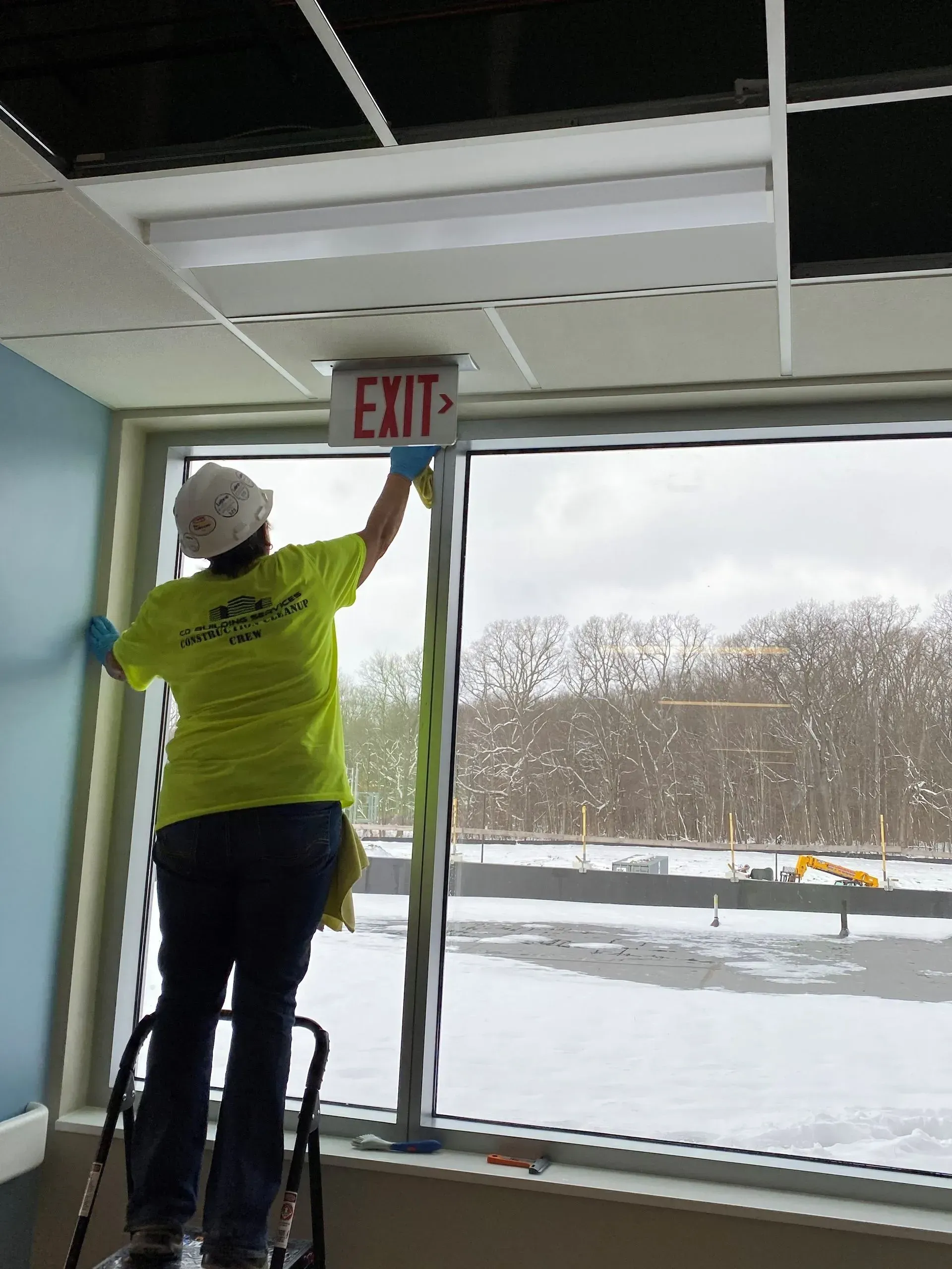 Worker in neon shirt adjusting an EXIT sign above a glass doorway.