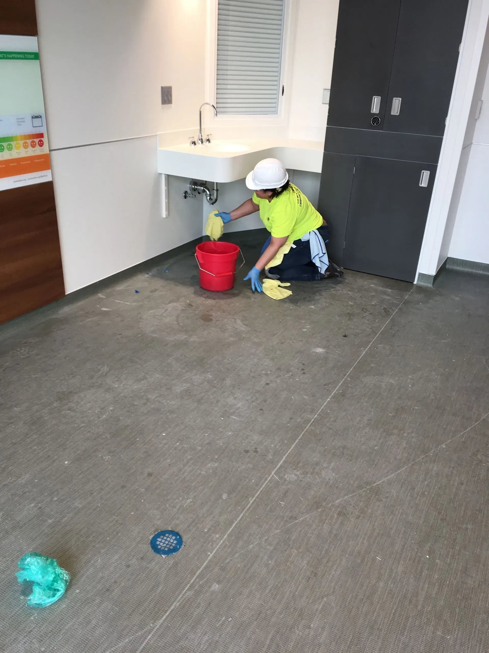 Worker in a hard hat crouches by a red bucket near a white sink in a hallway.