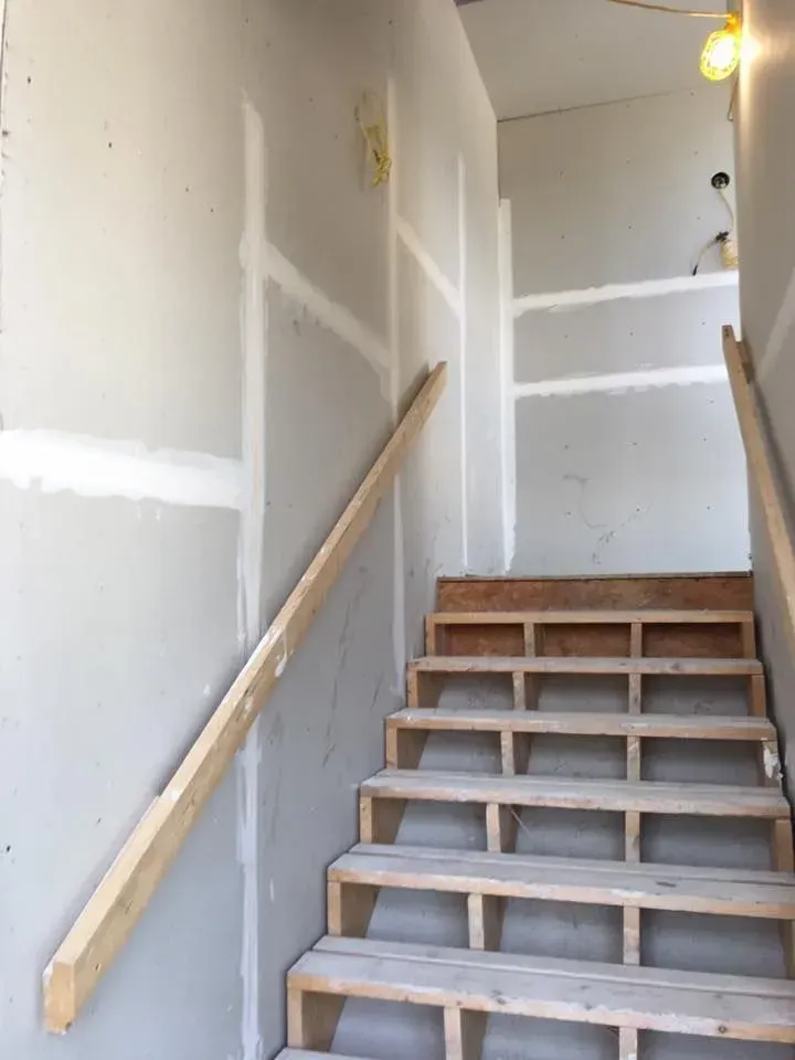Narrow unfinished stairwell with gray walls, wooden handrails, and steps leading upward to a lit landing