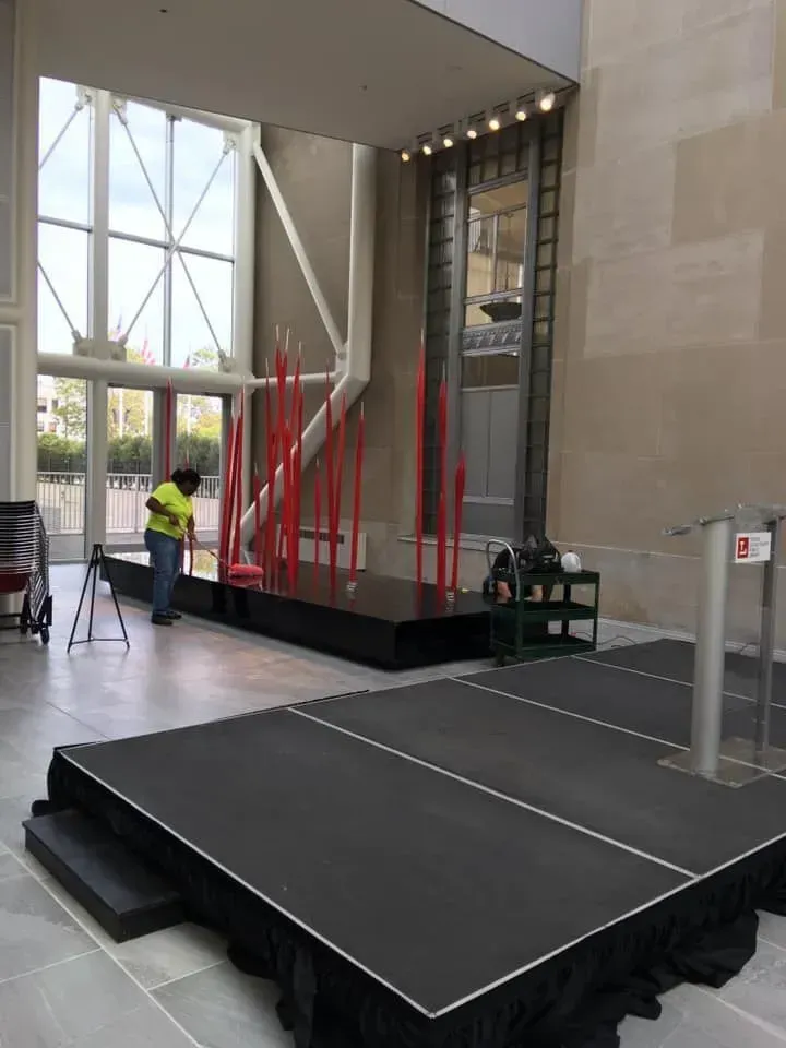Indoor stage with black platform, red cables, and a worker in a yellow safety vest near large windows