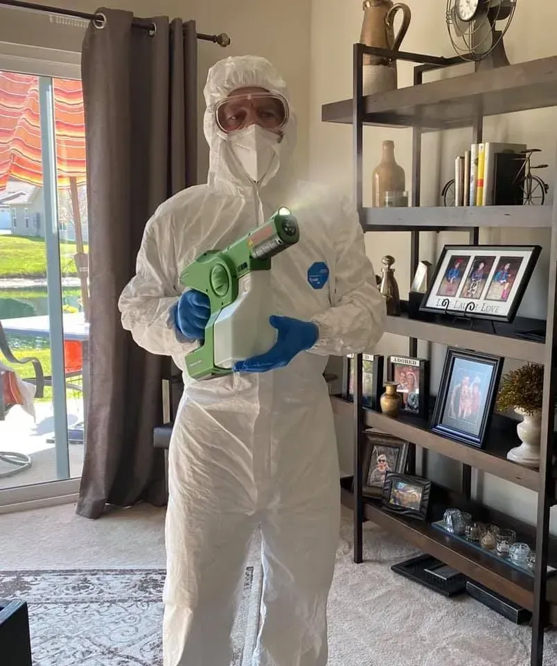 Person in white protective suit and mask holding a green spray gun inside a living room.