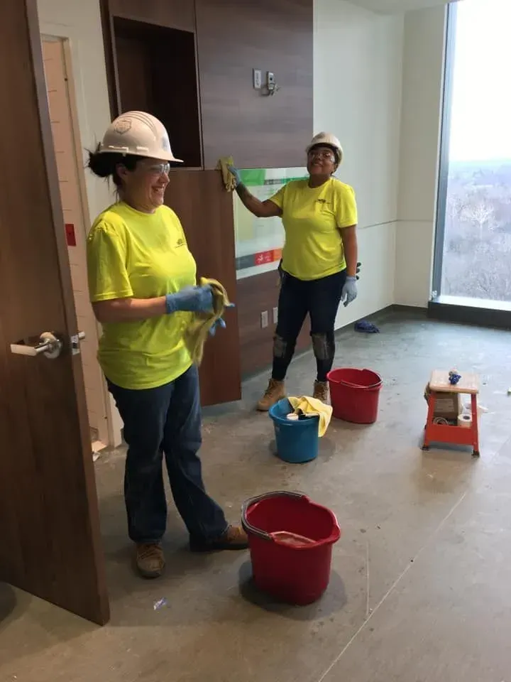 Two workers in yellow shirts and hard hats painting a room with buckets and tools on the floor