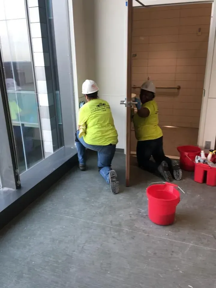 Two workers in yellow vests clean a hallway floor near windows and red buckets.