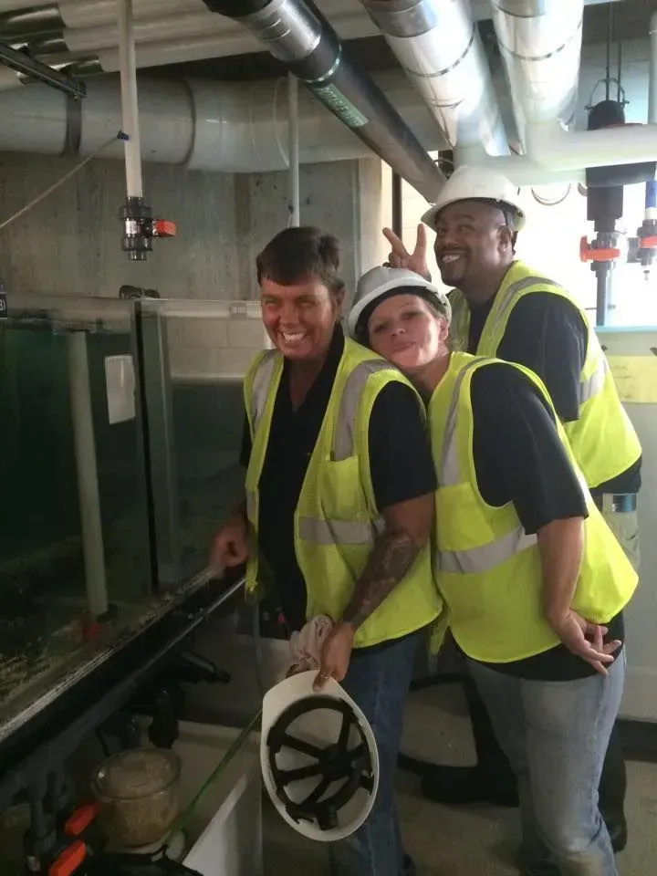 Three workers in yellow safety vests smiling in an industrial facility beside pipes and a valve wheel