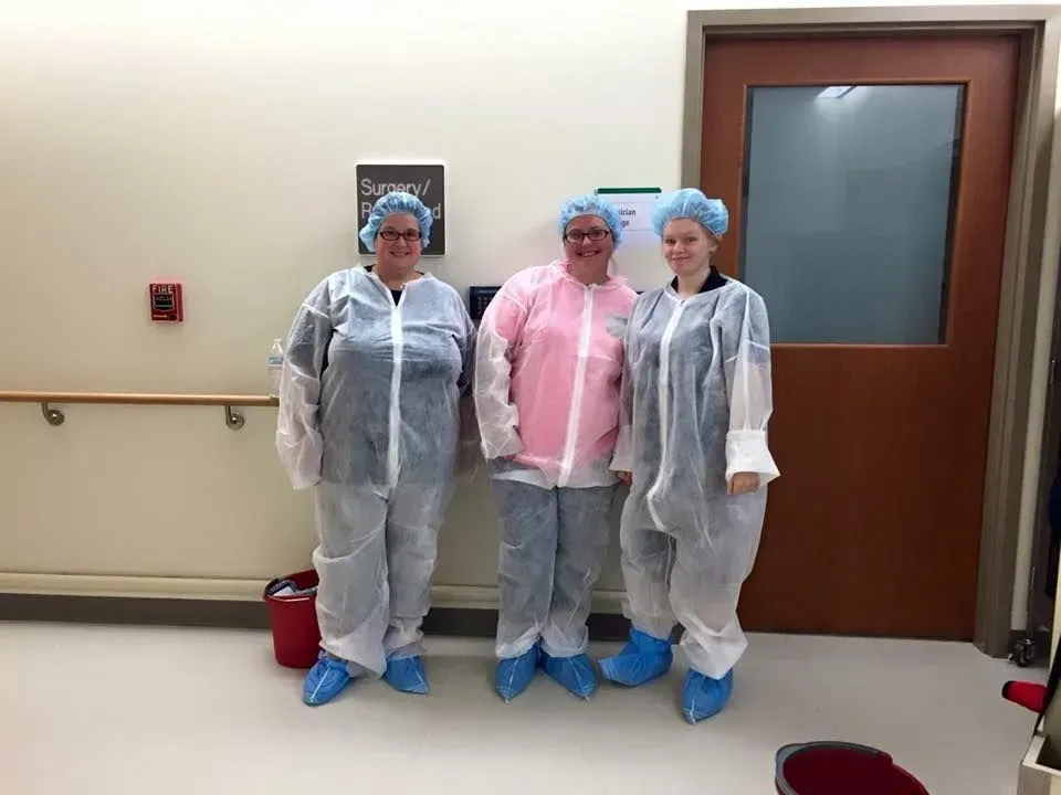 Three people in protective gowns and hair nets standing in a hospital hallway near a door.