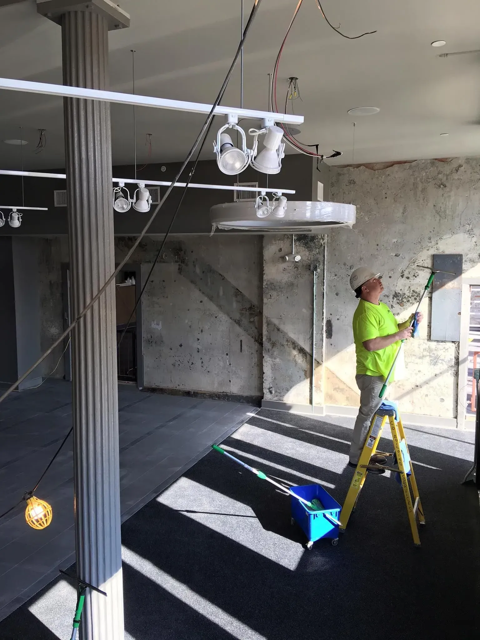 Person in neon vest painting a wall in an unfinished room with a ladder and hanging lights.