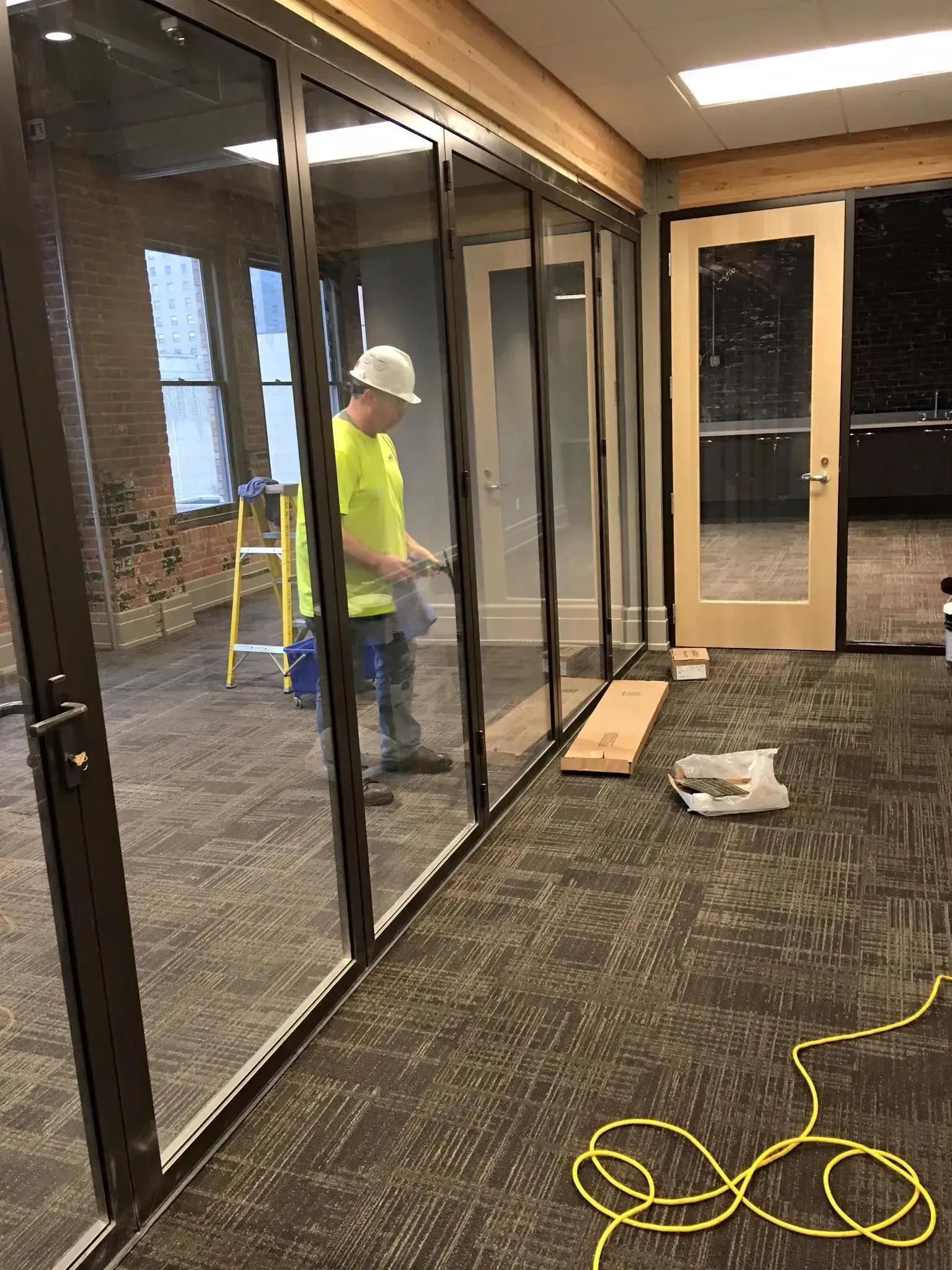Worker in a neon shirt installing glass panels in a modern hallway with tools and a ladder.