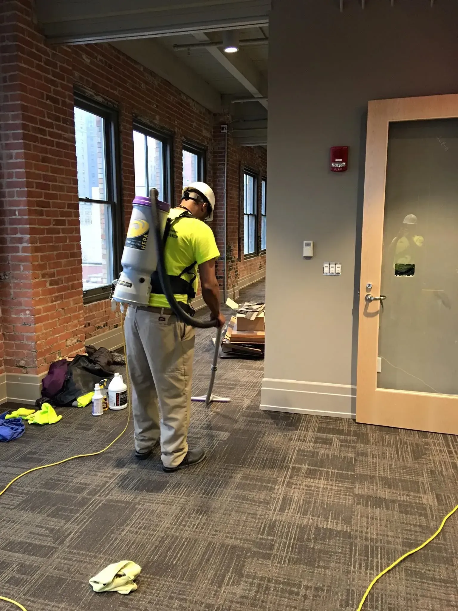 Worker in a yellow shirt and hard hat vacuuming debris in a hallway with brick walls and windows.