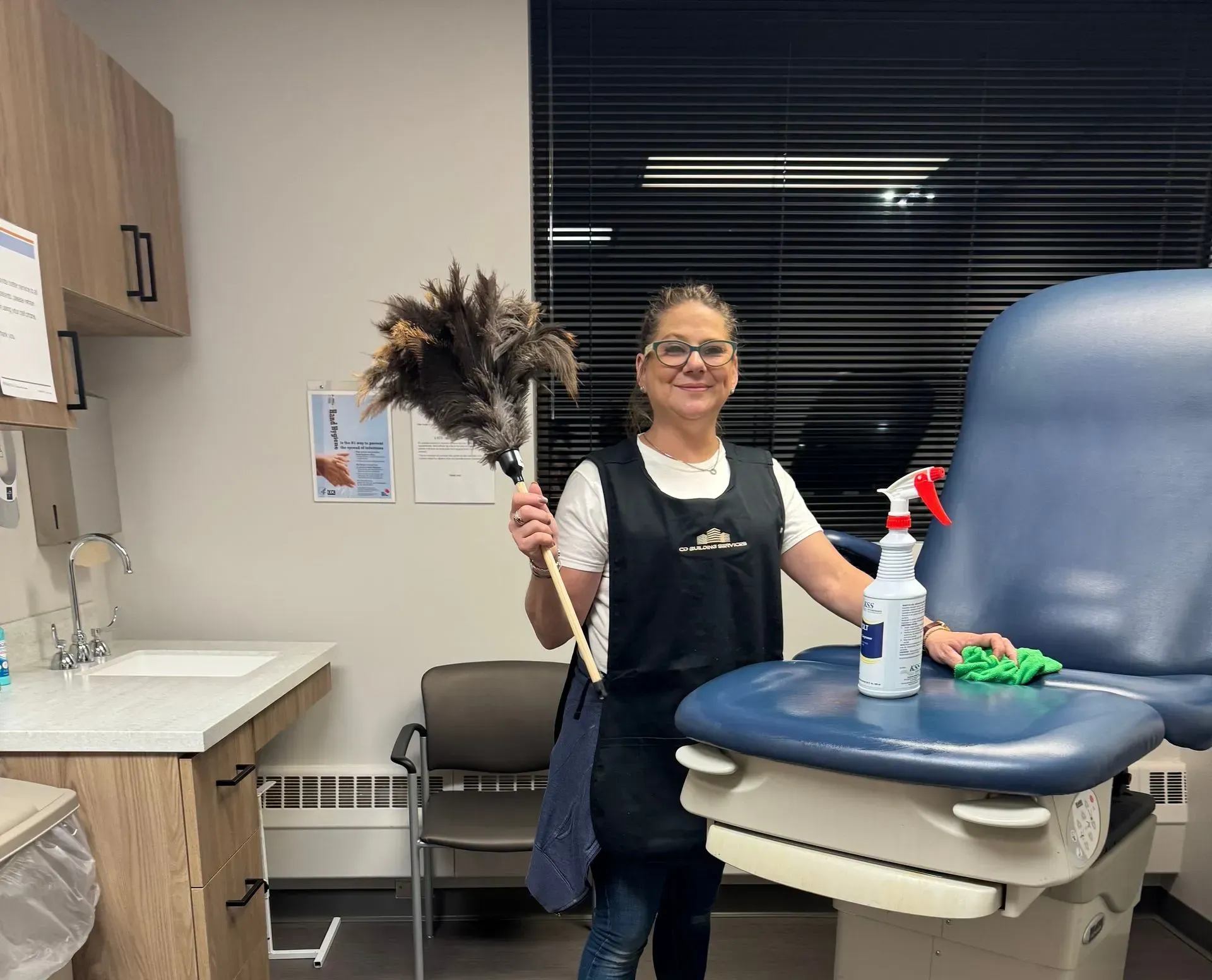 Person in a clinic exam room holding a feather duster beside a treatment chair and spray bottle.