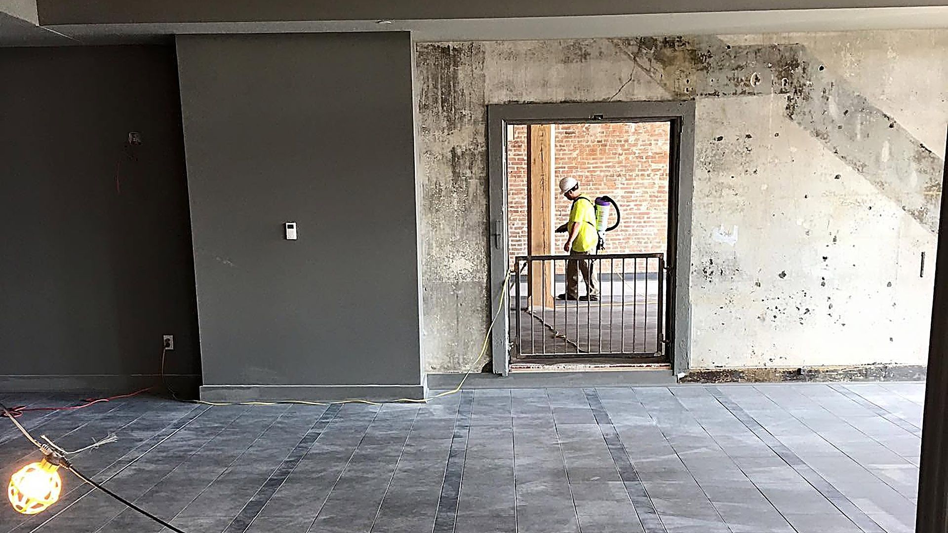 Empty unfinished room with tiled floor, concrete walls, and a doorway opening to a worker in yellow outside