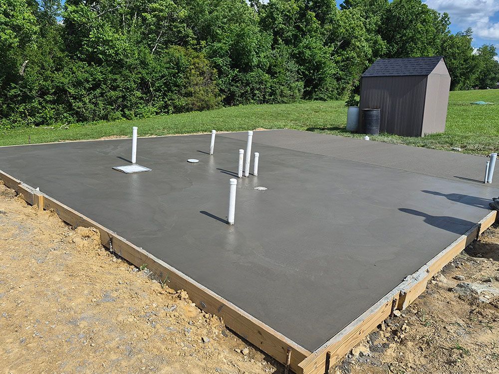 Concrete foundation slab with exposed plumbing pipes and a small shed in a grassy yard