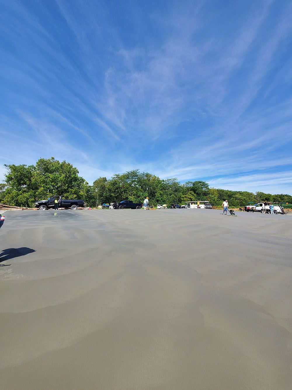 Wide sandy parking lot with scattered cars, green trees, and a bright blue sky with wispy clouds.
