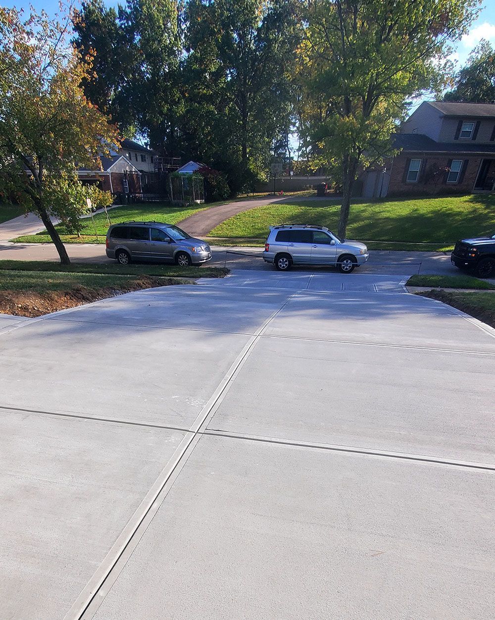 Driveway leading to a quiet street with parked cars and leafy trees in front of houses.