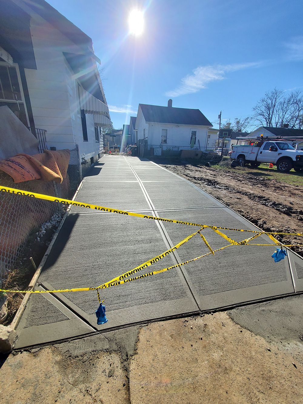 Caution-taped concrete walkway under construction beside houses, with trucks and dirt piles in bright sun