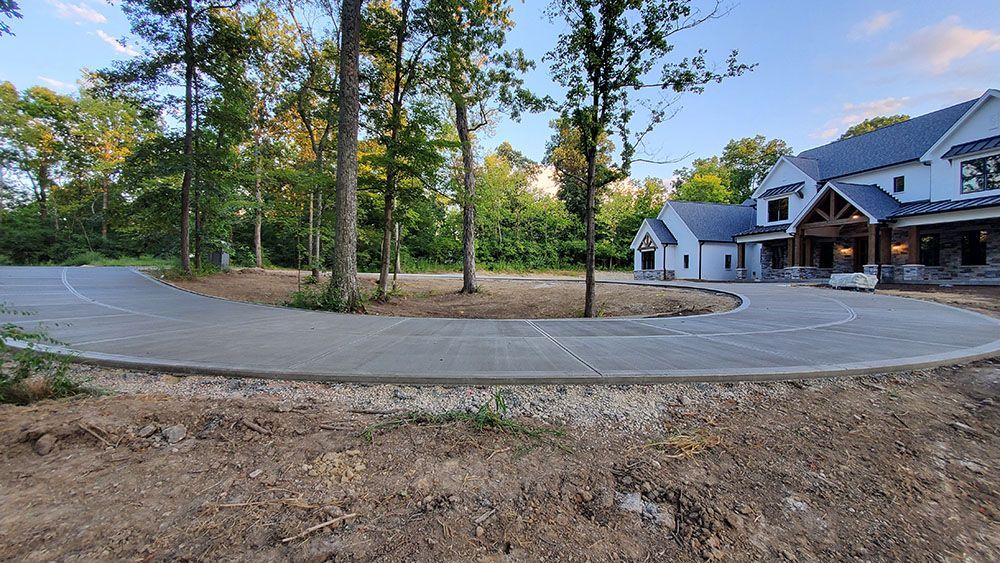 Curved driveway beside a wooded lot and a modern house under construction at dusk.