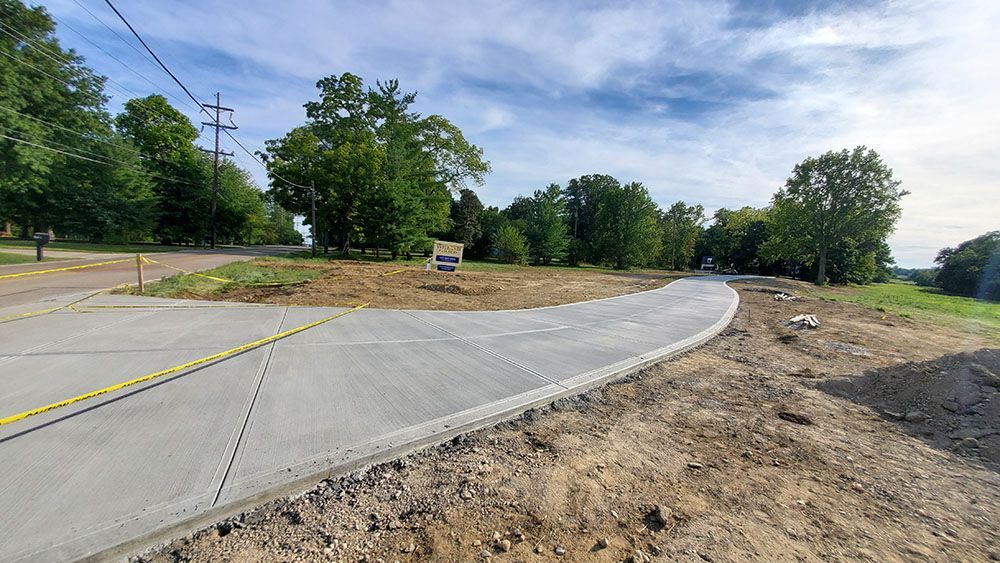 Curved concrete road under construction beside a dirt shoulder and trees on a sunny day