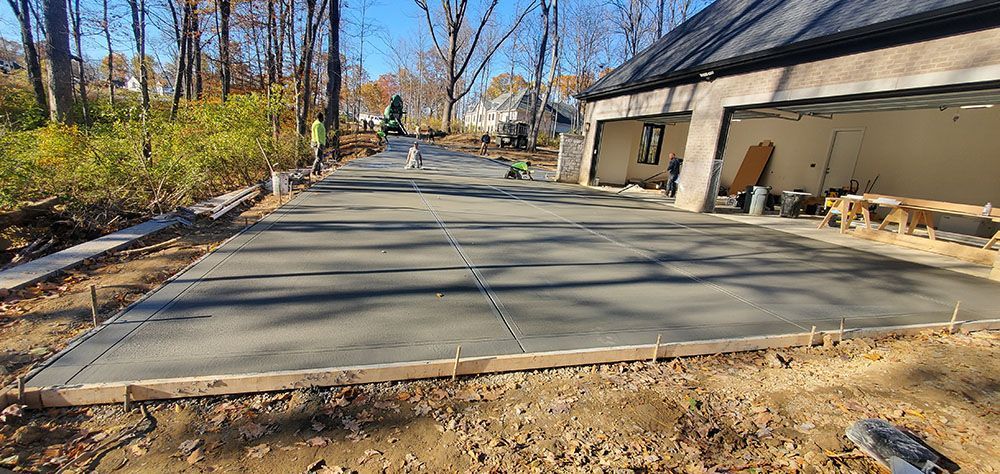 Concrete driveway under construction beside a house, with gravel edges and leafless trees nearby.