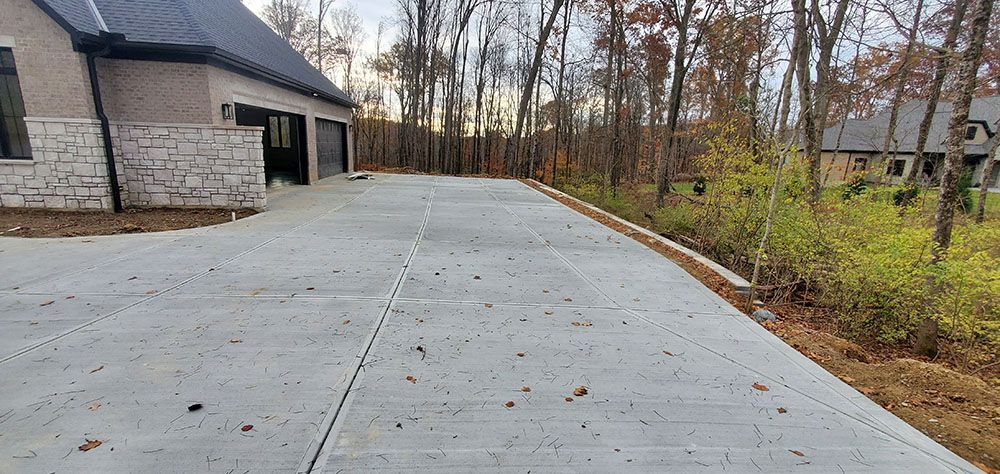 Concrete driveway beside a stone house, leading into a wooded yard in late autumn.