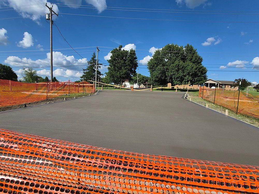 Paved area under construction with orange safety fencing, trees, and a blue sky.