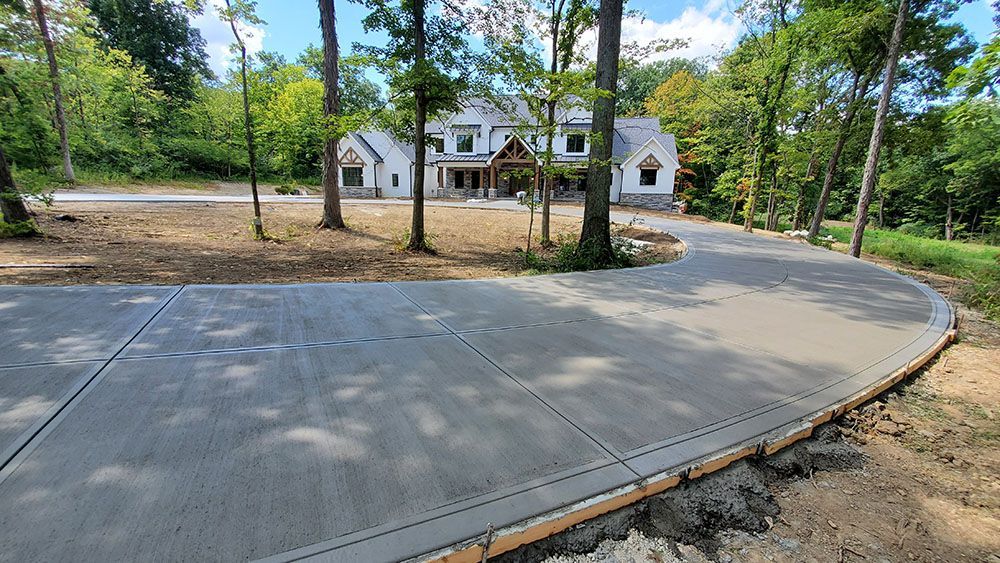 Curved concrete driveway leading to a white house amid trees in a wooded residential lot