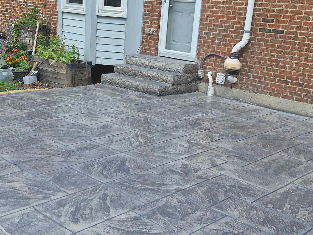 Gray stamped concrete patio beside a brick house with steps, flower pots, and a white downspout