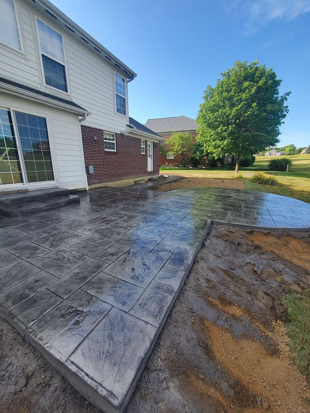 Wet concrete patio beside a house, with a tree and yard in the background.