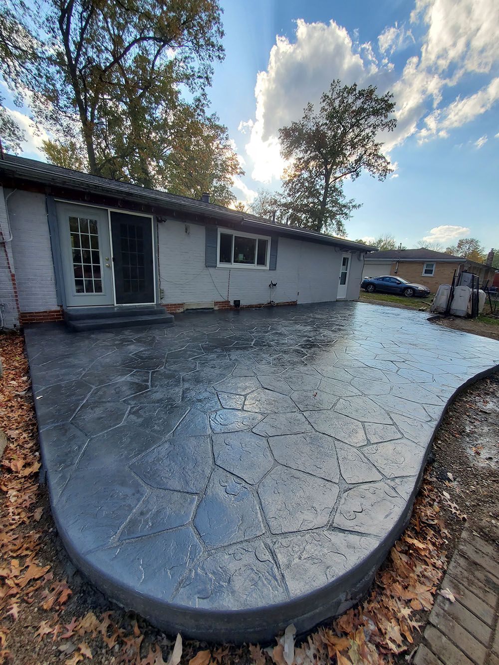 Curved stamped concrete patio beside a light gray house, with trees and parked cars in the background.