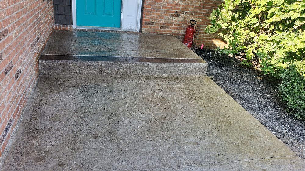 Concrete front porch with a small step leading to a teal door and brick wall, beside a gravel path and greenery