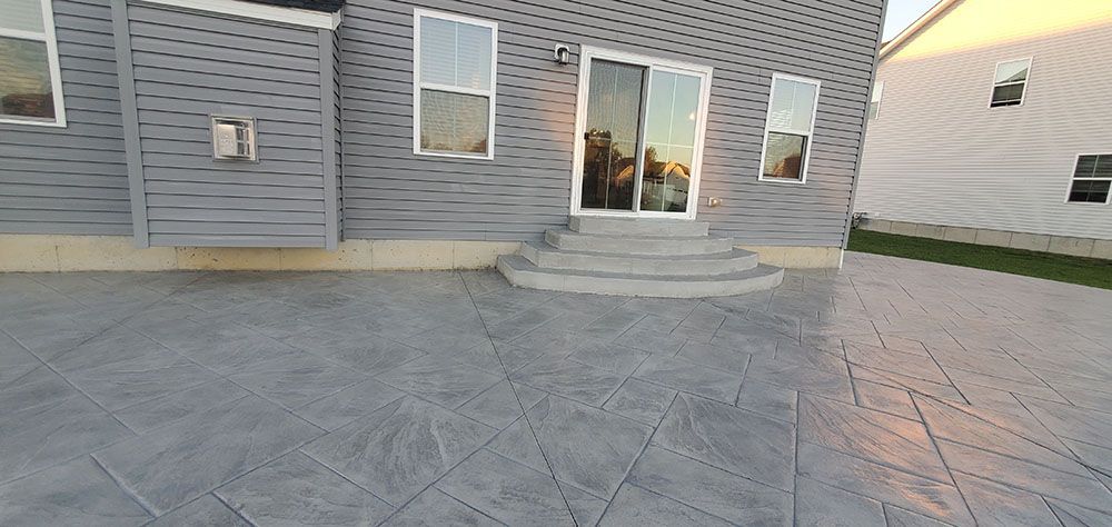 Patio with gray stone pavers leading to a house’s sliding glass door and steps, bordered by siding and grass.
