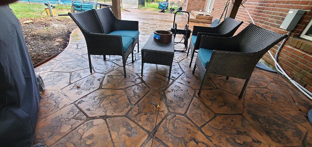 Rainy patio with wicker chairs and a small table on a stamped concrete floor, beside a brick wall.