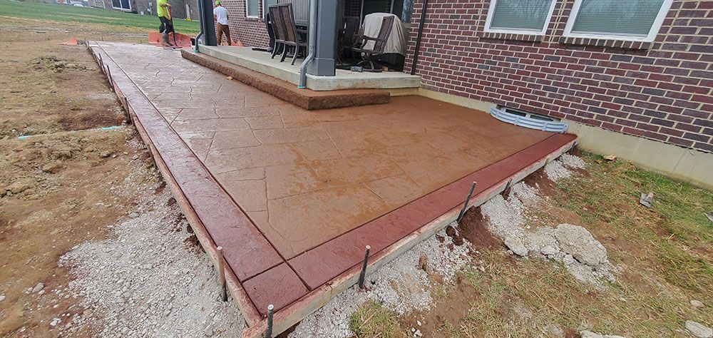 Brown stamped-concrete patio with uneven edge beside a brick house and exposed soil