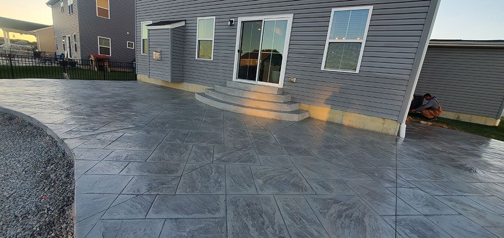 Stamped concrete patio beside gray house with sliding glass door and windows at sunset