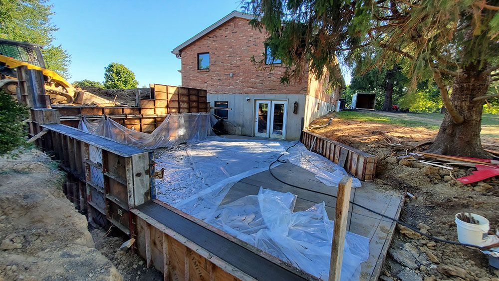 Backyard patio under construction beside a brick house, with covered ground and stacked lumber.