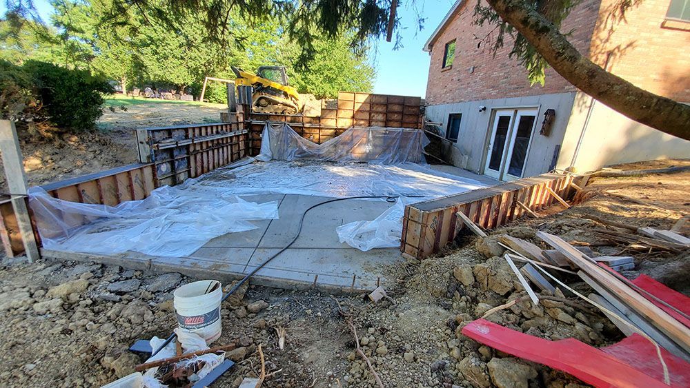 Patio construction site with rebar, concrete forms, tools, and a brick house in the background