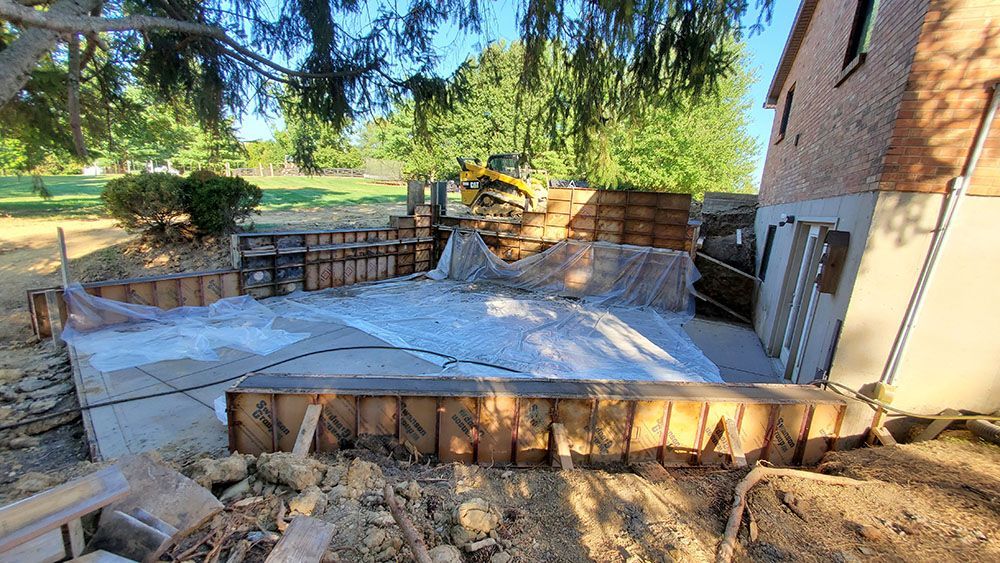 Empty concrete patio under construction beside a house, with retaining walls and lumber in a yard.