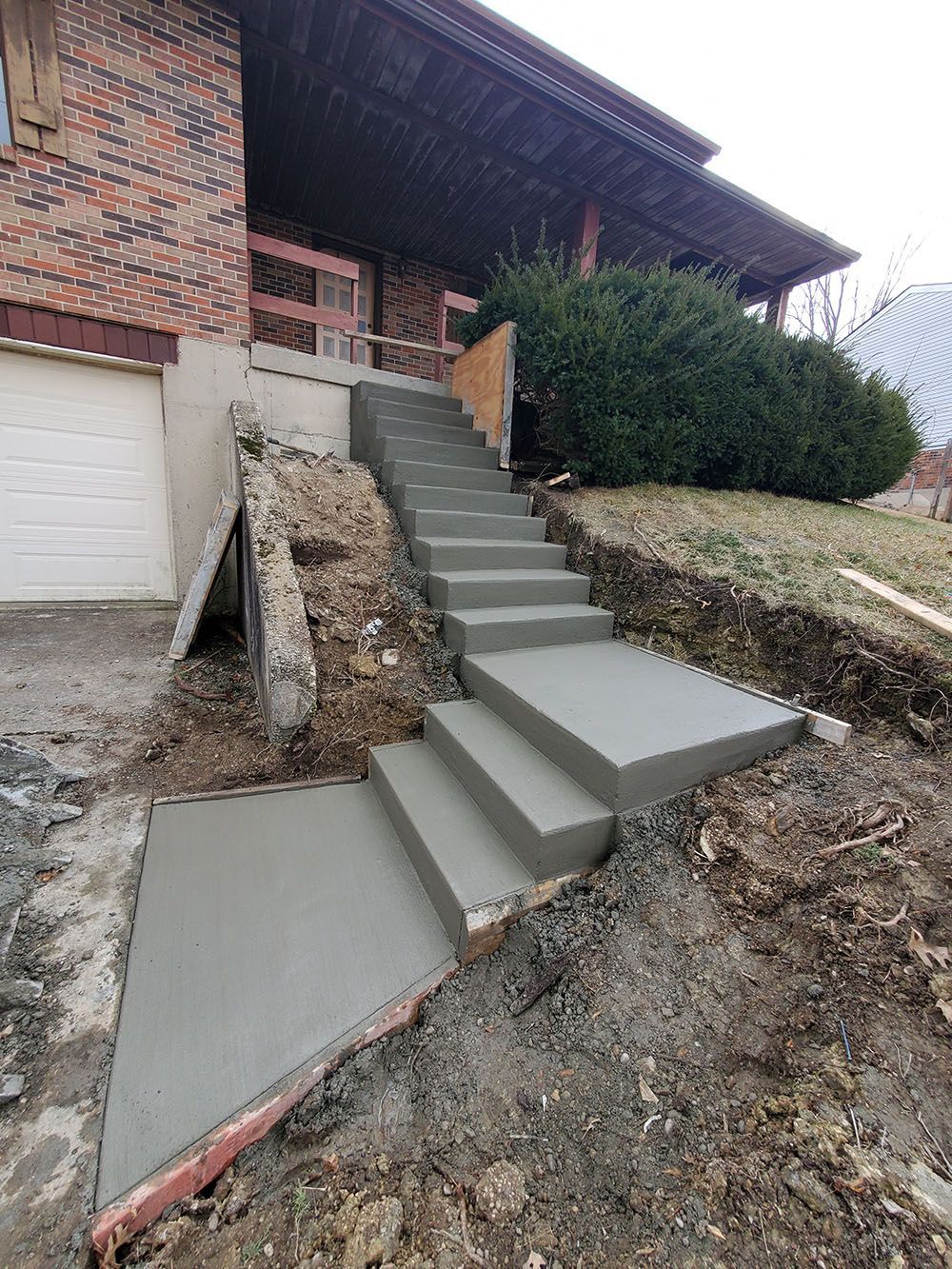 Concrete steps leading up a brick house entrance with a covered porch, bordered by shrubs and sloped dirt.