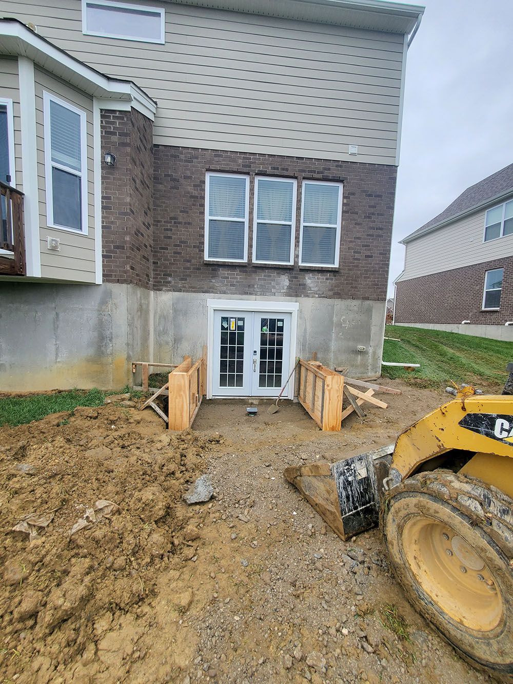 Backyard construction site with muddy ground, foundation walls, and a yellow excavator beside a house.